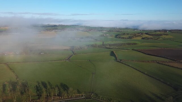 Drone footage of patchwork farmland and rural fields partially covered by drifting fog, revealing roads, trees and distant villages under clear blue sky. West Lothian, Scotland