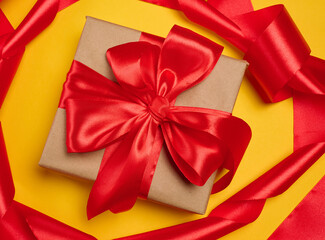 Gift box wrapped in craft paper and tied with a large bright red satin bow on a yellow background