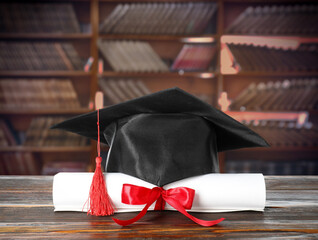 Rolled diploma tied with red ribbon and graduate cap on wooden table in library