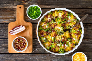 Naklejka premium Potato broccoli casserole - baked potatoes with broccoli, cheddar cheese and smoked bacon on wooden table. Top view