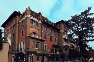 Obraz premium Fairy-tale villa in Quartiere Coppedè, Rome, by Gino Coppedè, with mosaic-tiled facade, arched entrance, tower with clock, balconies, and ornate details in warm tones under cloudy sky.