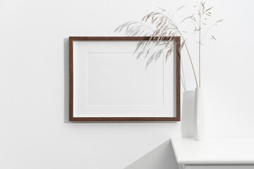 Blank horizontal dark wood frame on white wall next to tall white vase holding dried grass stems,...
