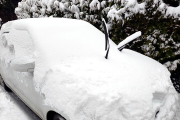 A white car under heavy snow with its windshield wipers raised, coated in powdery snow. A snow-covered car beside snowy hedge on parking lot