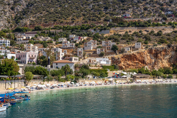Fototapeta premium Scenic view of Kalkan town beach with white umbrellas and turquoise water. Mediterranean coastal landscape with traditional houses on green hills, shot from marina pier. September, Turkey.