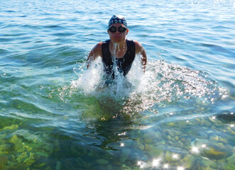 A young boy wearing swim goggles and a cap emerges from clear blue sea water, splashing energetically. Front view captures motion, summer light, and active seaside fun.