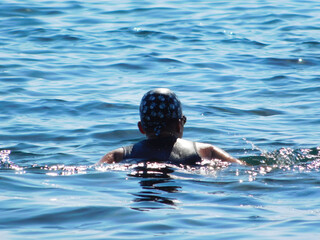 Wide-angle view of a young boy swimming freestyle in open blue sea on a sunny day, water splashing around him, conveying summer activity, fitness, freedom, and outdoor adventure