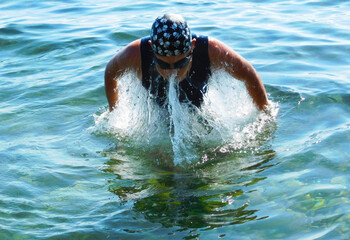 A young boy wearing swim goggles and a cap emerges from clear blue sea water, splashing energetically. Front view captures motion, summer light, and active seaside fun.