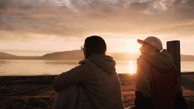 Two people sitting by the sea at sunset, enjoying a quiet moment and reflecting while watching the horizon. Silhouettes against warm evening light create a calm, thoughtful atmosphere of travel