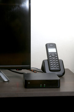 A vertical composition focusing on the workspace containing the 4K decoder, the base of a television, and a digital cordless phone.
