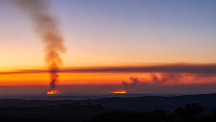 Obraz premium Dramatic sunset illuminates distant wildfires, with plumes of smoke rising against the vibrant orange and yellow sky, depicting an environmental crisis