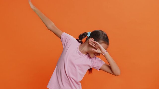 Trendy cheerful positive preteen child girl kid having fun dancing, moving to rhythm, dabbing raising hands, making dub dance winner celebrating victory gesture. Teenager children on orange background