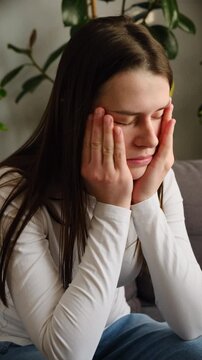 Tired unhealthy young woman sitting alone on couch at home with closed eyes, holding head with hand, suffering from strong sudden headache or migraine, throbbing pain concept