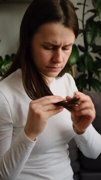 Frustrated worry young woman holding splitting ends, messy unbrushed dry hair and frizzy with disheveled hair sitting on couch at home. Damaged hair. Health care of beauty concept