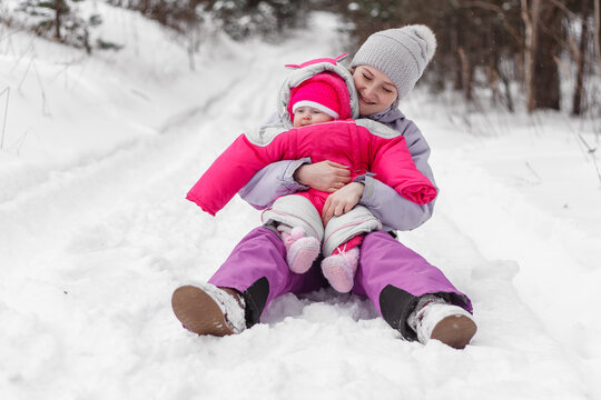 Woman sitting in snow, embracing baby, enjoying family time outdoors in winter park. Mother and child bonding during a cold weather walk.
