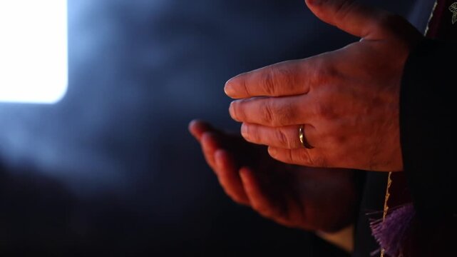Close up side view of the hands of a priest wearing traditional vestments making the sign of the cross during a religious ceremony with a dark background.

