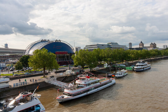 A wide panoramic view of the Rhine riverfront in Cologne, Germany, featuring the blue Musical Dome theater and various passenger boats docked at the embankment