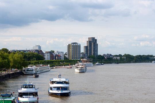 Scenic view of the Rhine river in Cologne, Germany, with several tourist cruise ships and passenger boats. Urban riverfront with modern apartment buildings under a cloudy sky