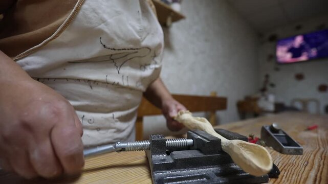 Close-up of artisan's hands tightening a metal vice to secure a carved wooden spoon on a workbench, preparing the utensil for further sanding and finishing work.

