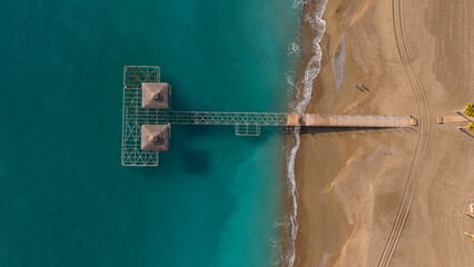 The symmetrical vertical pull of a modern wooden pier and thatched roofs over the deep blue sea. An aesthetic square representing luxury resorts and minimalist architecture.
