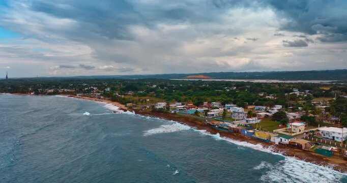 Raging waves hit the coast with row of buildings. Aerial perspective on the green cityscape in Puerto Rico under the grey cloudy sky.