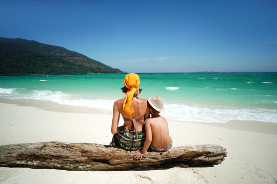 Rear view of a mother and child sitting together on a sandy tropical beach and looking at the turquoise sea. Summer vacation, family travel and lifestyle concept. 