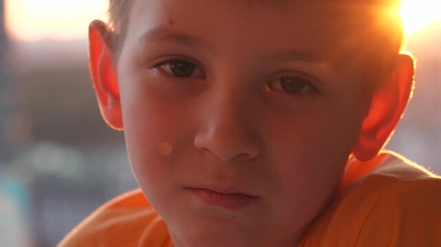 Pensive sad young boy face looking at camera and then tries smiling with warm sunset backlighting him. Atmospheric lens flare and an authentic emotional portrait of childhood innocence.