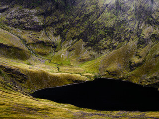 A dark, still tarn sits nestled in a valley among the rugged, green hills of National Park. Small streams trickle down the mountainside to feed the tarn waters.