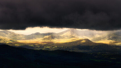 Sunlight filters through a gap in dark, stormy clouds, illuminating sections of mountainous terrain in National Park, creating a dramatic vista.