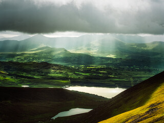 Sunlight streams through dark clouds, illuminating the green hills and fields of Killarney National Park. Lakes reflect the light in the foreground.