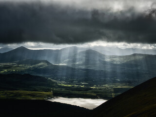 Sunlight breaks through storm clouds over the rolling hills and a lake in rural County Kerry, Ireland. The sunbeams illuminate the landscape during the daytime.