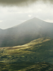 A high-angle view reveals the rolling, green landscape of County Kerry, Ireland. Sunlight illuminates the hills and valleys, while ominous clouds create a dramatic atmosphere above the mountains.