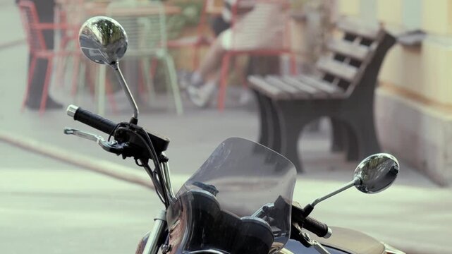 Closeup of motorcycle handlebars with mirrors and windscreen. Bench and chairs visible in blurred background
