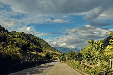Scenic rural road winds through green hills with trees and mountains under a partly cloudy sky, inviting a peaceful drive through nature and open countryside on a bright day