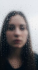 Blurred Face behind Glass. A blurred, out-of-focus portrait of a person behind a window with raindrops. The focus is on the water on the glass, making the person appear distant and hazy