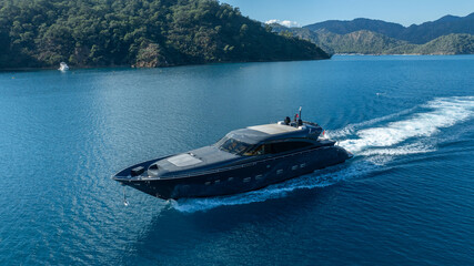 Obraz premium Luxury Yacht at Sea: The sleek, dark-hulled yacht carves through turquoise waters, a symbol of opulence and adventure, leaving a white wake against the stunning coastal backdrop.