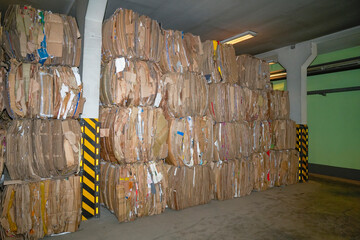 The packaging material has been flattened, pressed, and bound in preparation for recycling at a facility. Stacked compressed cardboard bales are seen in a brightly lit warehouse.