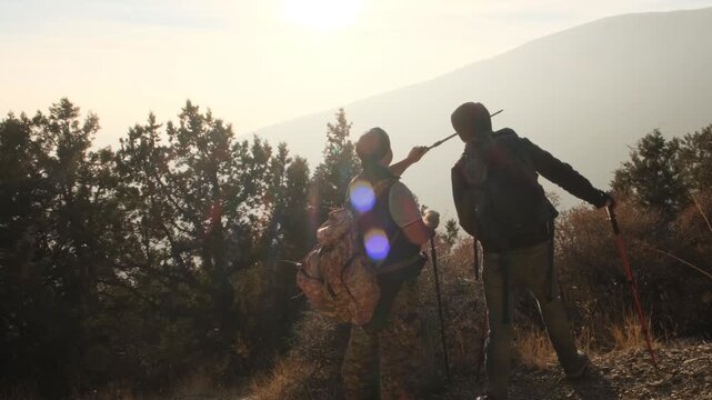 View from behind. Two traveling men stand facing the sunset and admire the mountain landscape pointing at something with a trekking pole. Enjoyment and happiness during travel. Hiking together.