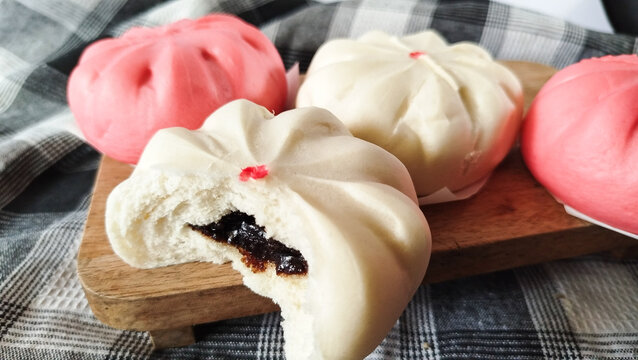 Sweet steamed buns known as bakpao presented on wooden cutting board. Soft white buns with chocolate paste filling, one opened, styled on checkered cloth background.