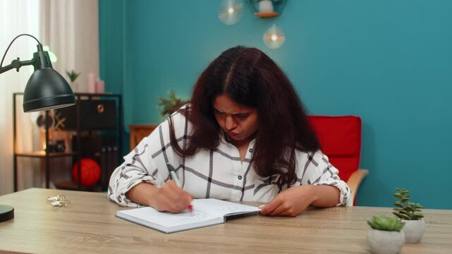 Indian woman at home writes idea in notebook crosses it out then retries showing determination to find solution. Arabian girl at table edits notes seeking creative answer with persistence and focus.