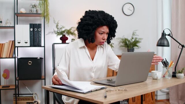 African American businesswoman at home office sees laptop crash glitch panics tossing papers urgently. Freelancer girl at table hits power button reboot hoping data safe returns and virus cleared soon