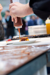 Fototapeta premium Chef drizzling sauce onto plated dish at culinary event or restaurant. Professional food preparation and fine dining presentation for hospitality, catering, and food service content.