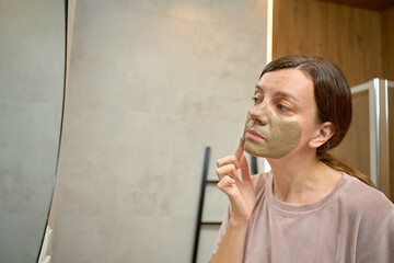Woman standing in bathroom and applying clay face mask while looking at mirror