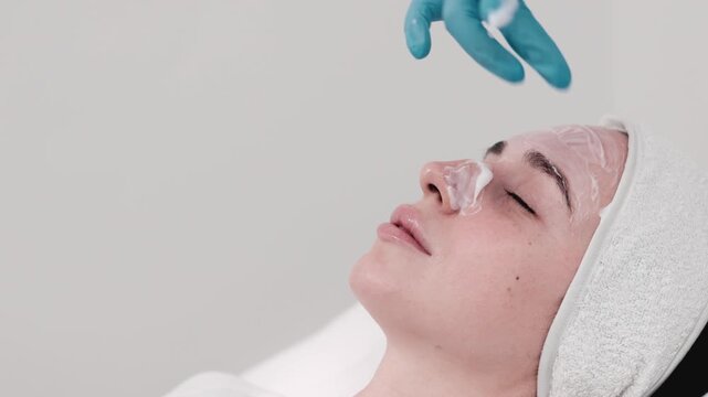 Female aesthetician in blue gloves applies facial treatment cream to a woman's face, demonstrating a professional skincare procedure and careful product application. Concept Professional skincare, Bea