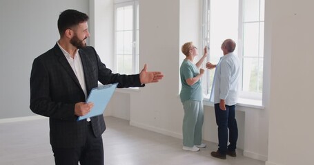 Realtor and real estate agent guide senior couple through a bright interior at an apartment viewing for realty, noting large windows with a clipboard. Elderly family tours an empty house
