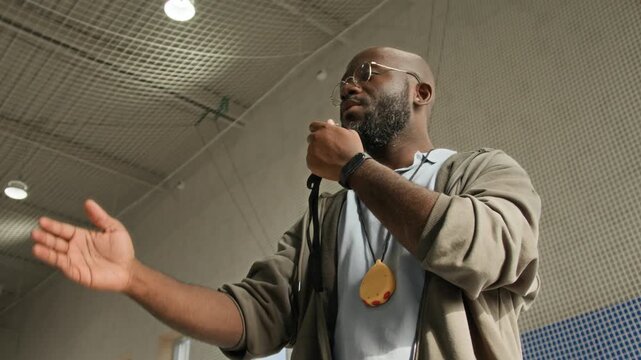 Low angle shot of Black male coach blowing whistle and gesturing actively while giving commands during sports match in indoor gym, copy space