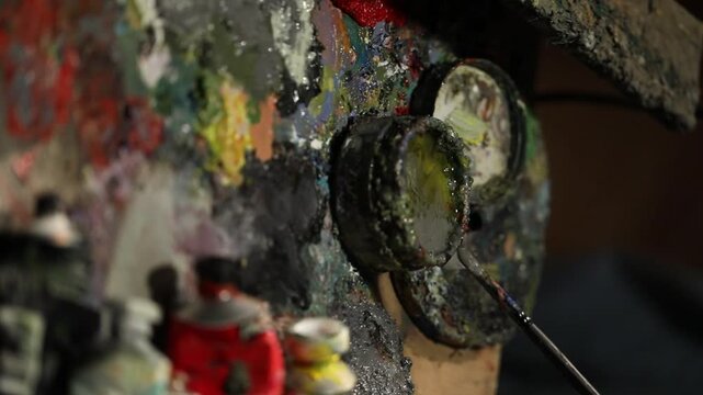 Close-up of artist's hand dipping paintbrush into solvent cup on colorful wooden palette. Detailed shot of creative painting process in art studio environment with oil paints.

