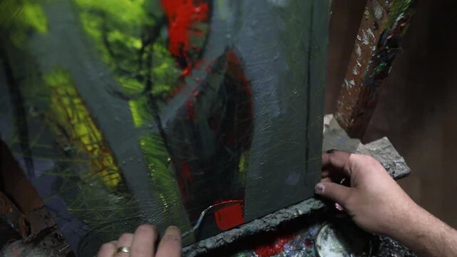 Close-up of artist's hand dipping paintbrush into solvent cup on colorful wooden palette. Detailed shot of creative painting process in art studio environment with oil paints.

