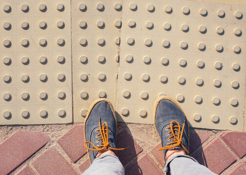 Women's feet in sneakers stand on a tactile tile. The road for the blind.
