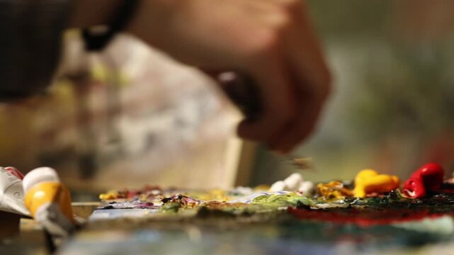 Extreme close-up of artist's fingers crushing a blob of yellow oil paint on a messy, colorful palette with brushes nearby in a dark atmospheric art studio.
