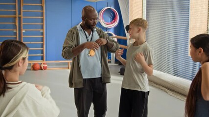 Waist up shot of Black man teaching PE class explaining shoulder rolls exercise to group of children and correcting technique